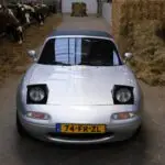 Een vooraanzicht van een grijze Mazda Miata in een stal met koeien