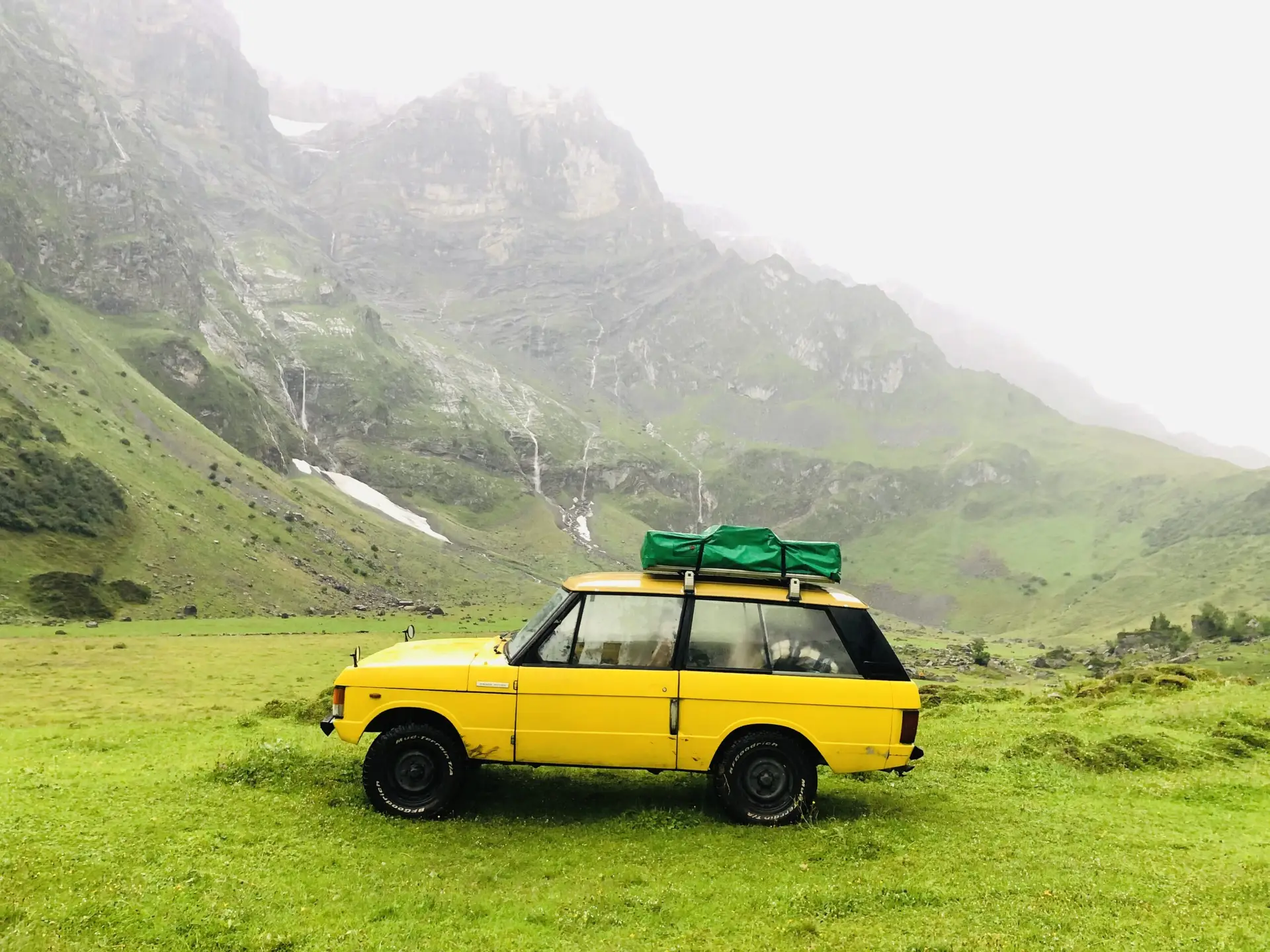 A yellow Range Rover Suffix C with a rooftent on top standing between the mountains